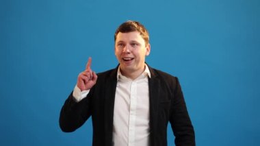Inspired business man in jacket pointing finger up with open mouth, showing Eureka gesture, solution, idea, inspiration, answer on blue studio background