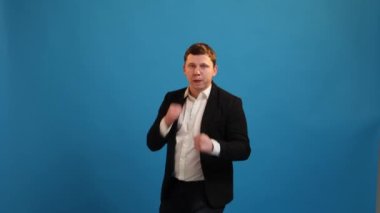 Businessman standing with boxing gesture, punching to camera, provoking fight or ready to self-defense, wearing suit on blue studio background