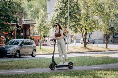 Happy smiling woman traveler is riding her electro scooter in city parkland with dog Welsh Corgi Pembroke in a special backpack