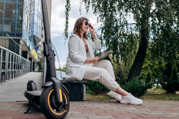 Beyaz takım elbiseli, güneş gözlüklü, gülümseyen iş kadını modern ofis binasının yanında elektrikli scooterıyla dijital tablet üzerinde çalışıyor. 