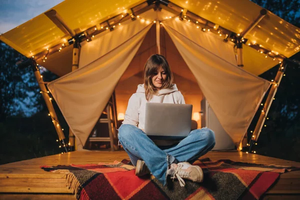 Happy Woman freelancer using a laptop on a cozy glamping tent in a summer evening. Luxury camping tent for outdoor holiday and vacation. Lifestyle concept