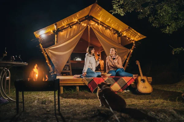 Two Smiling female friends drinking wine and eating fruits sitting in cozy glamping tent in autumn evening bonfire. Luxury camping tent for outdoor holiday and vacation. Lifestyle concept