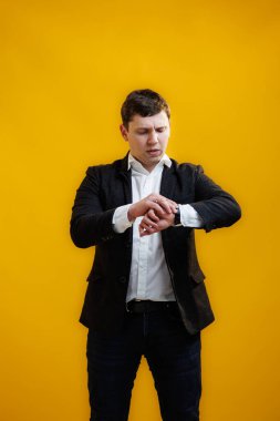Young businessman in suit checking time on his wrist watch standing on yellow studio background. Concept of punctuality