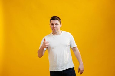 Young handsome man wearing white t-shirt over yellow background approving doing positive gesture with hand, thumbs up smiling and happy for success. Winner gesture 