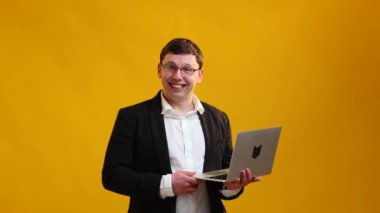 Smiling businessman speaker in glasses showing on laptop while talking on camera over yellow studio background. Business concept