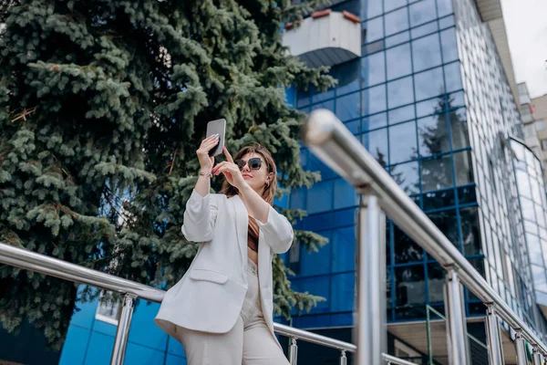 Smiling business woman in white suit and sunglasses making selfie using phone during break standing near modern office building