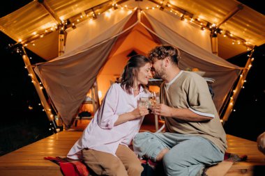 Happy couple relaxing in glamping on summer evening and drinking wine near cozy bonfire. Luxury camping tent for outdoor recreation and recreation. Lifestyle concept