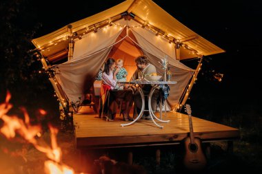 Happy family with lovely baby have dinner and spend time together in glamping on summer evening near cozy bonfire. Luxury camping tent for outdoor recreation and recreation. Lifestyle concept