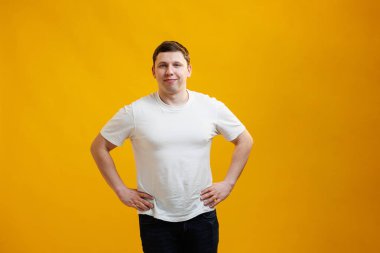 Young handsome man wearing white t-shirt with happy face smiling and looking at the camera over yellow studio background. Positive person