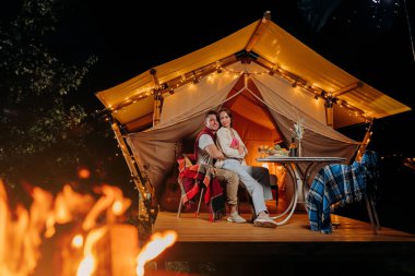 Happy lovely couple relaxing in glamping on summer evening and drinking wine near cozy bonfire. Luxury camping tent for outdoor recreation and recreation. Lifestyle concept