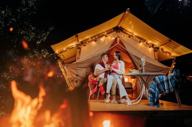 Happy lovely couple relaxing in glamping on summer evening and drinking wine near cozy bonfire. Luxury camping tent for outdoor recreation and recreation. Lifestyle concept