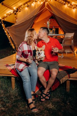 Happy lovely couple with Welsh Corgi Pembroke dog relaxing in glamping on evening and drinking wine near cozy bonfire. Luxury camping tent for outdoor recreation and recreation. Lifestyle concept