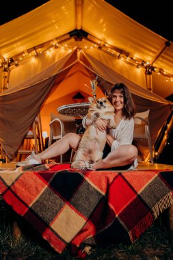 Happy young woman with her Welsh Corgi Pembroke dog relaxing in glamping on summer evening near cozy bonfire. Luxury camping tent for outdoor recreation and recreation. Lifestyle concept