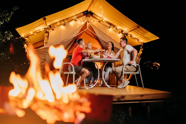 stock image Happy group of friends relaxing in glamping and drinking wine on summer evening near cozy bonfire. Luxury camping tent for outdoor recreation and recreation. Lifestyle concept