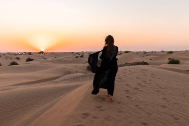 Beautiful mysterious woman in traditional arabic black long dress stands in the desert on sunset
