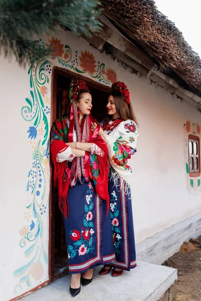Two Ukrainian women in traditional ethnic clothing and floral red wreath on background of decorated hut. Ukrainian national embroidered dress call vyshyvanka. Pray for Ukraine
