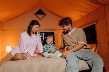 Happy family with lovely baby playing and spend time together in cozy glamping on summer evening while lying on bed. Luxury camping tent for outdoor recreation and recreation