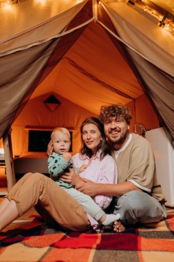 Happy family with lovely baby playing and spend time together in glamping on summer evening. Luxury camping tent for outdoor recreation and recreation. Lifestyle concept