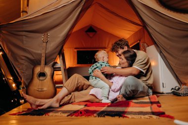 Happy family with lovely baby playing and spend time together in glamping on summer evening. Luxury camping tent for outdoor recreation and recreation. Lifestyle concept