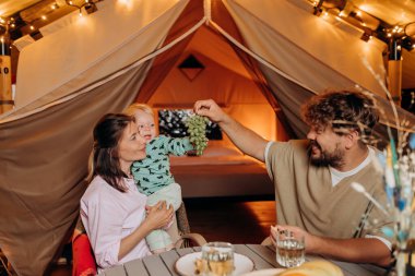 Happy family with lovely baby have dinner and spend time together in glamping on summer evening near cozy bonfire. Luxury camping tent for outdoor recreation and recreation. Lifestyle concept