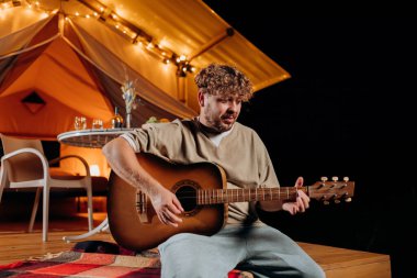 Handsome young smiling man playing guitar sitting in cozy glamping tent in summer evening bonfire. Luxury camping tent for outdoor holiday and vacation. Lifestyle concept