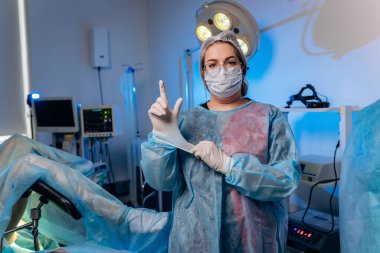 Female proctologist wearing medical uniform is posing showing fingers and smiling in hospital before operation clipart