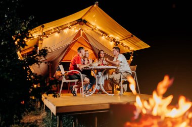 Happy group of friends relaxing in glamping and drinking wine on summer evening near cozy bonfire. Luxury camping tent for outdoor recreation and recreation. Lifestyle concept