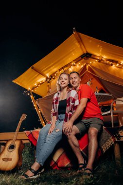 Happy lovely couple relaxing in glamping on evening near cozy bonfire. Luxury camping tent for outdoor recreation and recreation. Lifestyle concept