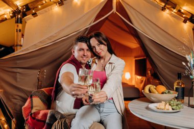 Happy lovely couple relaxing in glamping on summer evening and drinking wine near cozy bonfire. Luxury camping tent for outdoor recreation and recreation. Lifestyle concept