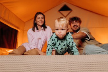 Happy family with lovely baby playing and spend time together in cozy glamping on summer evening while lying on bed. Luxury camping tent for outdoor recreation and recreation