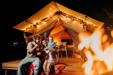 Happy family relaxing and spend time together in glamping on summer evening and playing guitar near cozy bonfire. Luxury camping tent for outdoor recreation and recreation. Lifestyle concept