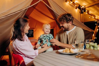 Happy family with lovely baby have dinner and spend time together in glamping on summer evening near cozy bonfire. Luxury camping tent for outdoor recreation and recreation. Lifestyle concept