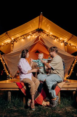 Happy family with lovely baby relaxing and spend time together in glamping on summer evening near cozy bonfire. Luxury camping tent for outdoor recreation and recreation. Lifestyle concept