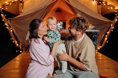 Mutlu bir aile, güzel bir bebeği rahatlatıyor ve yaz akşamları birlikte glamping yaparak sıcacık şenlik ateşinin yanında vakit geçiriyorlar. Açık hava eğlencesi ve dinlenme için lüks kamp çadırı. Yaşam tarzı kavramı