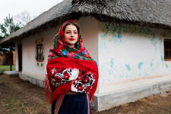 Charming woman in traditional ukrainian handkerchief, necklace and embroidered dress standing at background of decorated hut. Ukraine, style, folk, ethnic culture