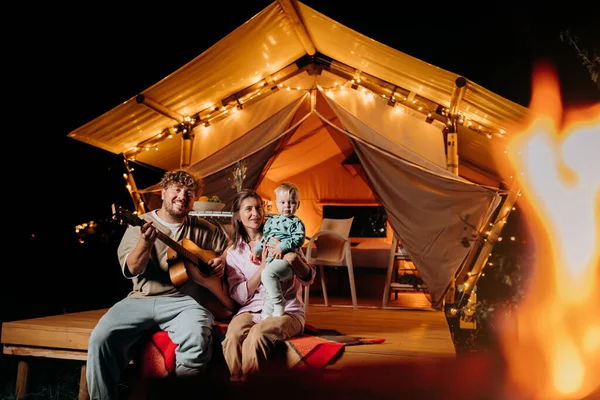 stock image Happy family relaxing and spend time together in glamping on summer evening and playing guitar near cozy bonfire. Luxury camping tent for outdoor recreation and recreation. Lifestyle concept