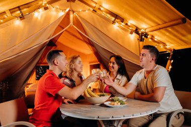 Happy group of friends relaxing in glamping and drinking wine on summer evening near cozy bonfire. Luxury camping tent for outdoor recreation and recreation. Lifestyle concept