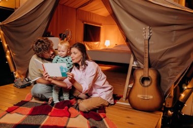 Güzel bir bebeği olan mutlu bir aile yaz akşamları glamping 'de birlikte kitap okuyor. Açık hava eğlencesi ve dinlenme için lüks kamp çadırı. Yaşam tarzı kavramı