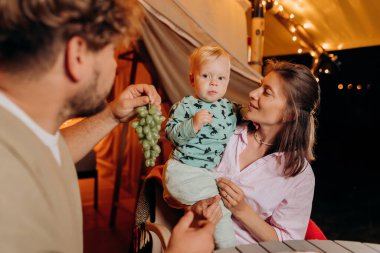 Happy family with lovely baby have dinner and spend time together in glamping on summer evening near cozy bonfire. Luxury camping tent for outdoor recreation and recreation. Lifestyle concept