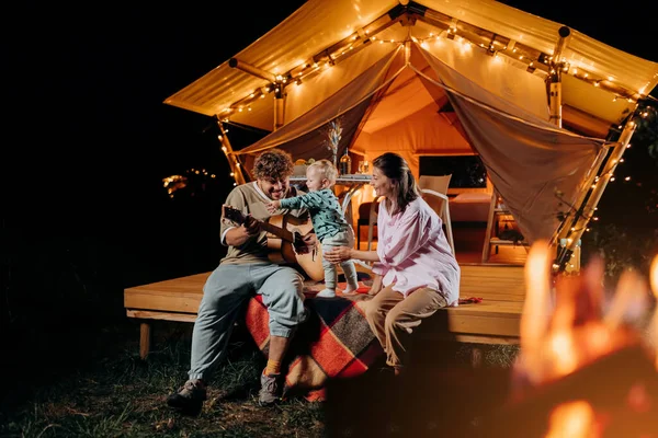 Happy family relaxing and spend time together in glamping on summer evening and playing guitar near cozy bonfire. Luxury camping tent for outdoor recreation and recreation. Lifestyle concept