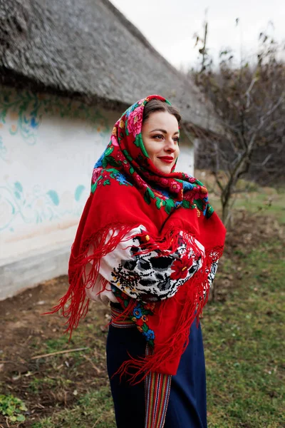 Charming woman in traditional ukrainian handkerchief, necklace and embroidered dress standing at background of decorated hut with thatched roof. Ukraine, style, folk, ethnic culture