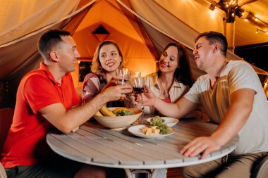 Happy group of friends relaxing in glamping and drinking wine on summer evening near cozy bonfire. Luxury camping tent for outdoor recreation and recreation. Lifestyle concept