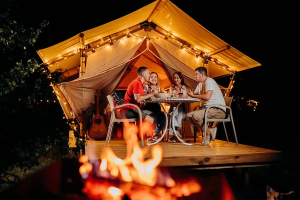 stock image Happy group of friends relaxing in glamping and drinking wine on summer evening near cozy bonfire. Luxury camping tent for outdoor recreation and recreation. Lifestyle concept