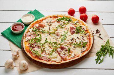 Salamlı, rukkola ve mantarlı taze pişmiş pizza. Ahşap zemin üzerinde domates, sos ve otlarla servis ediliyor. Yemek teslimatı konsepti. Restoran menüsü