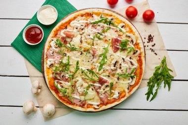 Salamlı, rukkola ve mantarlı taze pişmiş pizza. Ahşap zemin üzerinde domates, sos ve otlarla servis ediliyor. Yemek teslimatı konsepti. Restoran menüsü. Üst görünüm