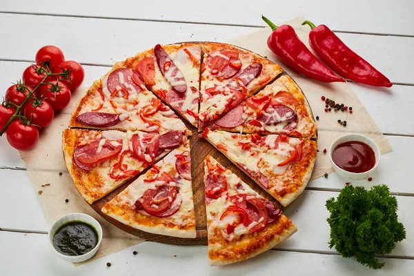 stock image Freshly baked pizza with smoked sausages, red pepper and tomatoes served on wooden background with sauce and herbs. Food delivery concept. Restaurant menu