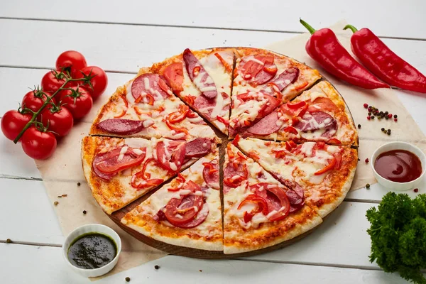 stock image Freshly baked pizza with smoked sausages, red pepper and tomatoes served on wooden background with sauce and herbs. Food delivery concept. Restaurant menu