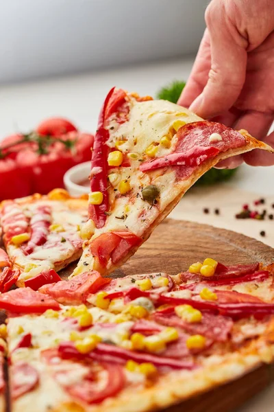The human hand takes Freshly baked tasty pepperoni pizza with salami, mozzarella cheese, corn and pepper served on wooden background with tomatoes, sauce and herbs. Food delivery concept. Restaurant menu
