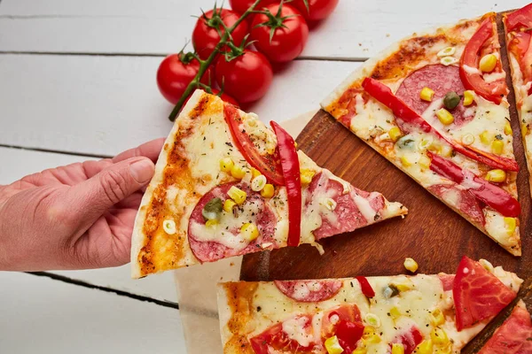 İnsan eli, salamlı, mozzarella peynirli, mısır ve biberli taze pişmiş pepperoni pizzayı ahşap arka planda domates, sos ve bitkilerle servis eder. Yemek teslimatı konsepti. Restoran menüsü