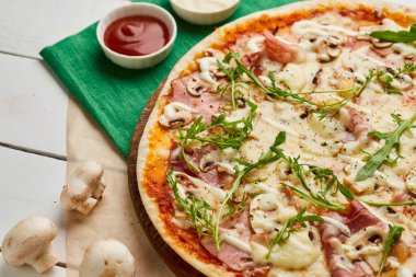 Salamlı, rukkola ve mantarlı taze pişmiş pizza. Ahşap zemin üzerinde domates, sos ve otlarla servis ediliyor. Yemek teslimatı konsepti. Restoran menüsü